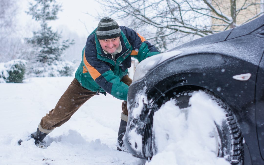 A person wearing heavy winter clothing attempts to push a car stuck in deep snow on a snowy day, underscoring the importance of seasonal tire changes and winter automotive services at Scott's Auto.