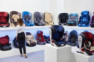 A woman that is looking at various car seats in a store. There is one row on a bottom shelf of car seats and a row above that. 