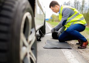 A man that has gotten a flat tire and has pulled off to the side of the road. He has put on a reflector vest to be seen by other cars and a car mat for the ground to kneel on. He is starting to raise the car up with a car jack.