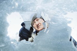 A view from inside the car looking at a man outside the scar that is scraping the ice off of a windshield with a white ice scraper. 