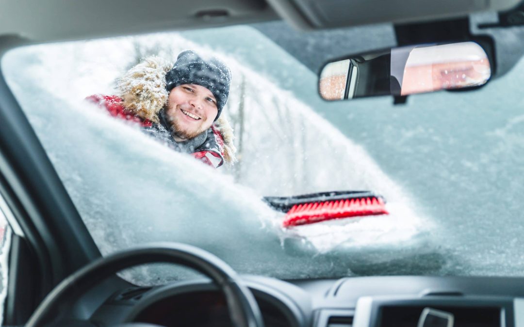 View from inside a car looking out at a smiling man in winter clothing clearing a snow-covered windshield with a red brush tool, emphasizing the importance of working wiper blades, heating system maintenance, and winter driving preparation at Scott's Auto.