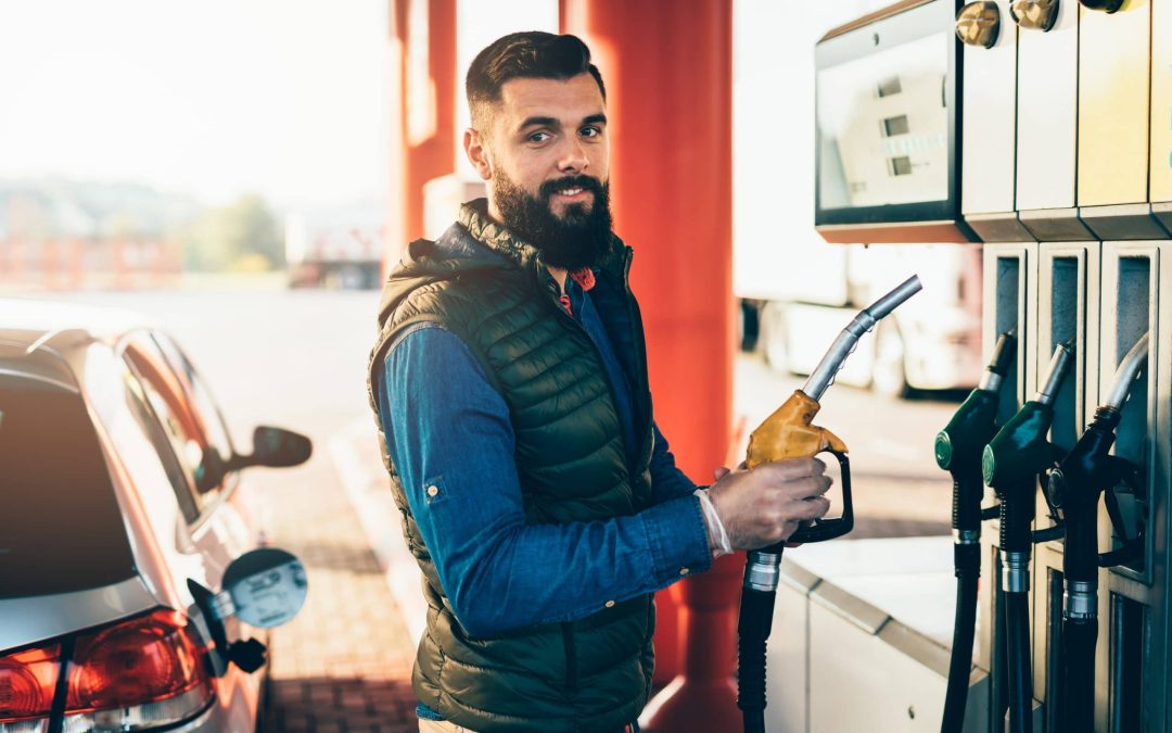 A bearded man holding a fuel nozzle at a gas station pump next to his car, emphasizing the importance of fuel system cleaning and regular vehicle maintenance available at Scott's Auto.