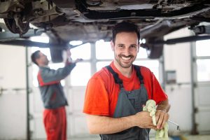 Car professionals that are working underneath a car. One of them is smiling at the camera and cleaning a dipstick. 