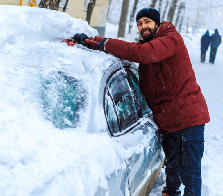 Driver using a snow brush and scraper to clear heavy snow off car roof during winter weather prep at Scott's Fort Collins Auto.