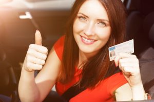 A beautiful brunette woman that is giving the camera a thumbs-up and holding her new license in her other hand. 
