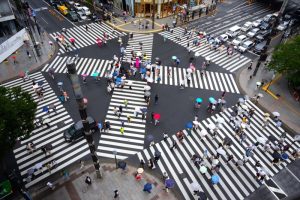 A very large intersection in a huge city. There are 6 different crosswalks in the intersection with crowds of people walking as traffic waits for them.