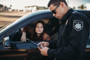 A police officer that is looking at a paper as a woman driver is waiting in a car next to him. The woman was pulled over due to a traffic violation.