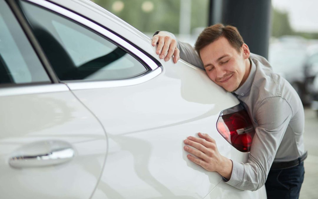 A smiling man affectionately hugging the rear panel of a clean white car, demonstrating a driver's appreciation for a well-maintained vehicle thanks to high-quality car care and routine automotive repair services from Scott's Auto.