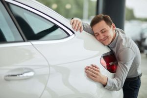 A smiling man affectionately hugging the rear panel of a clean white car, demonstrating a driver's appreciation for a well-maintained vehicle thanks to high-quality car care and routine automotive repair services from Scott's Auto.